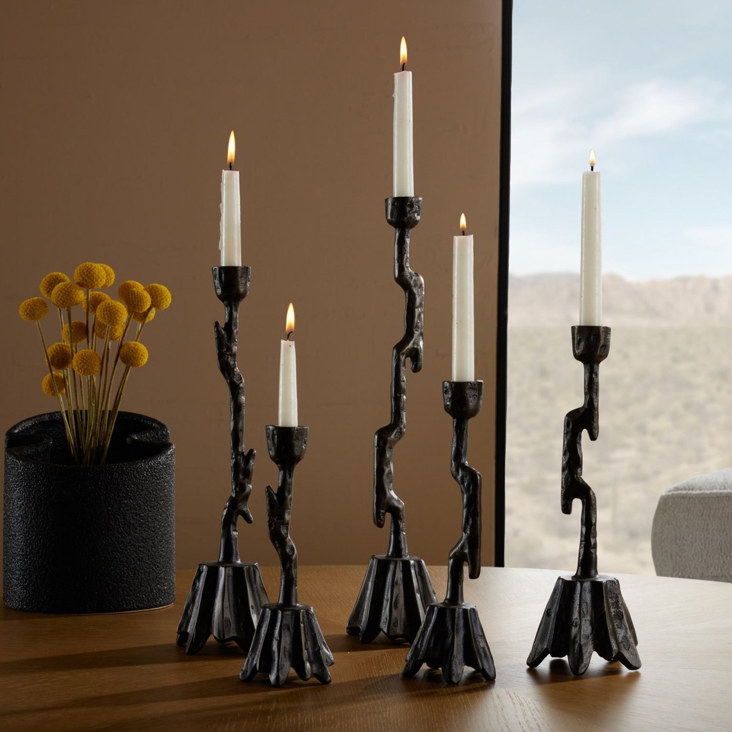 Set of five Brandt Candlesticks styled with lit candles, paired with a dark vase holding orange flowers.