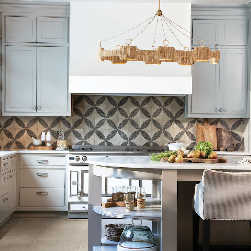 Raffi Large LED Linear Chandelier styled in a transitional kitchen above the island, accented with fruit and vegetables, light blue cabinetry, a white range hood, and a beautifully detailed backsplash.