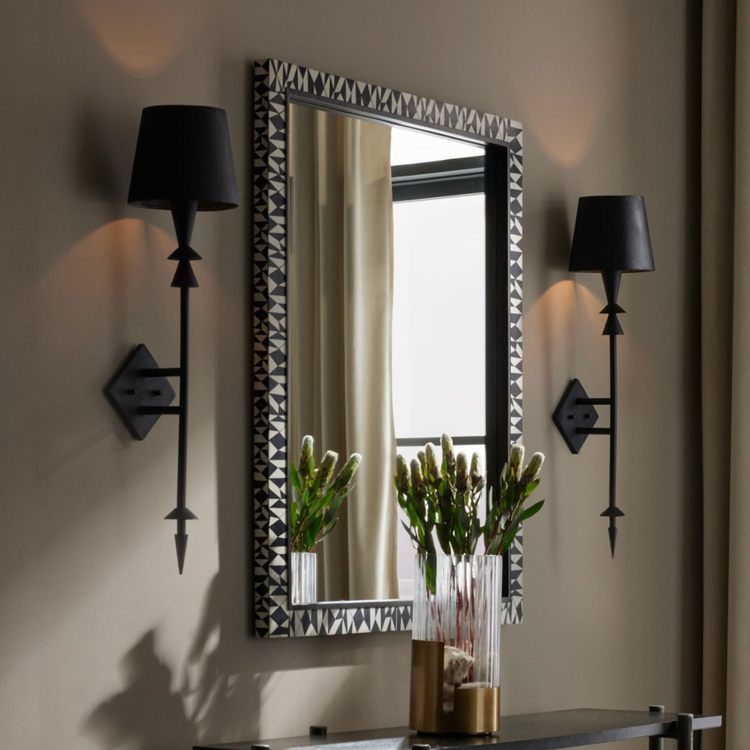 Decorative wall mirror with a patterned frame, flanked by two black wall sconces, styled above a console table with a glass vase of greenery.