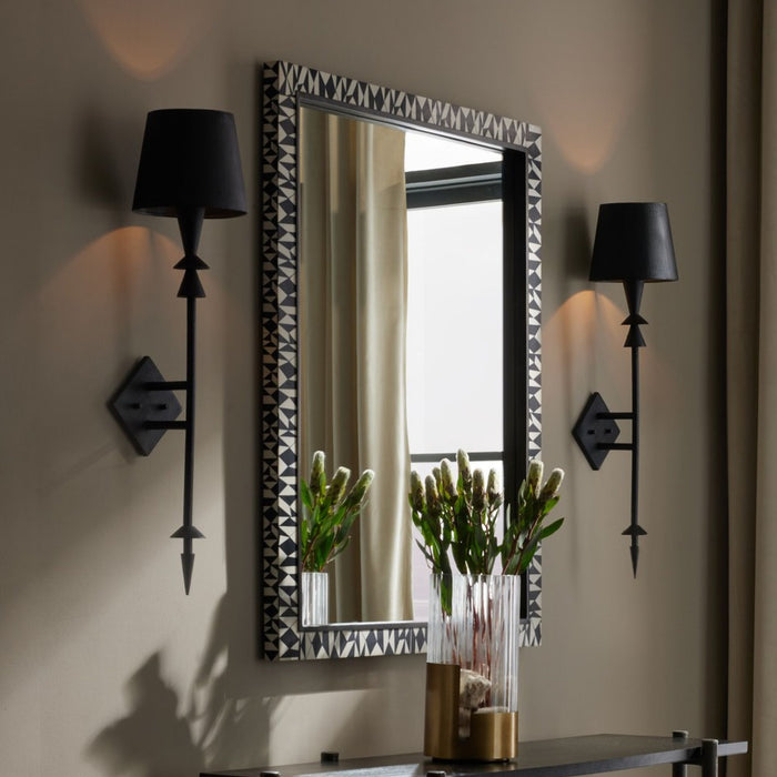 Decorative wall mirror with a patterned frame, flanked by two black wall sconces, styled above a console table with a glass vase of greenery.