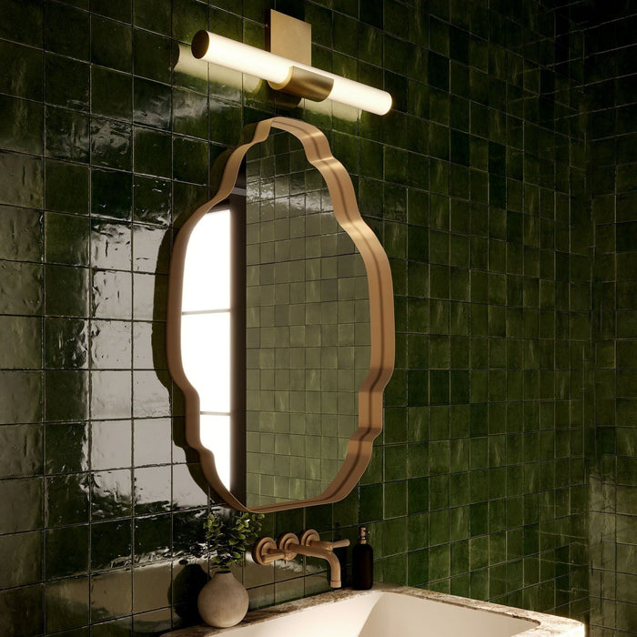 Apollo LED Wall Vanity in a vintage brass finish installed above a brass-framed vanity mirror and sink, set against deep green tiled bathroom walls.