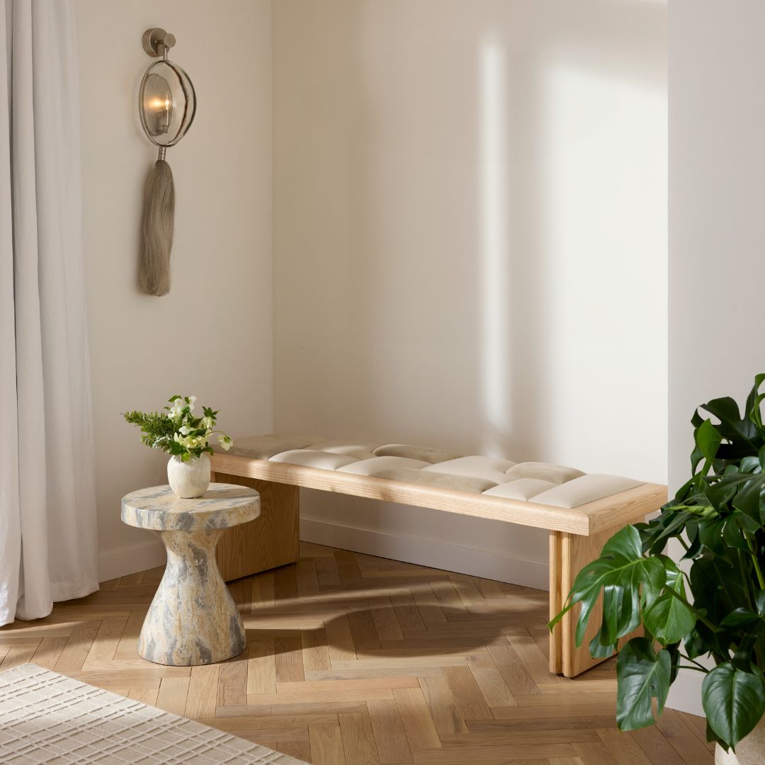 Fairfield Bench styled in an entryway alongside a green plant, set against light wood flooring, with a wall sconce to the left and a side table topped with a plant.