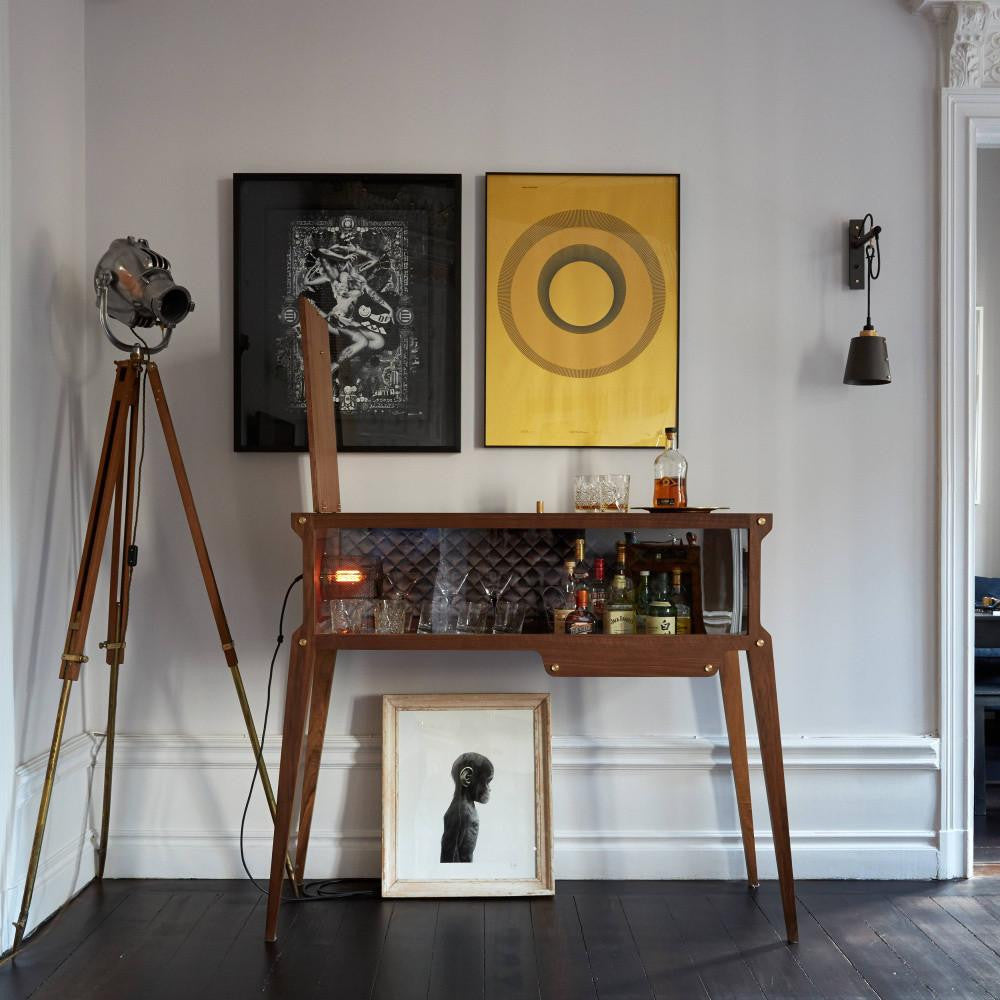 Rockstar Bar styled against a wall lined with spirits and glassware, with two paintings above, a wall sconce to the right, and an industrial floor lamp to the left.