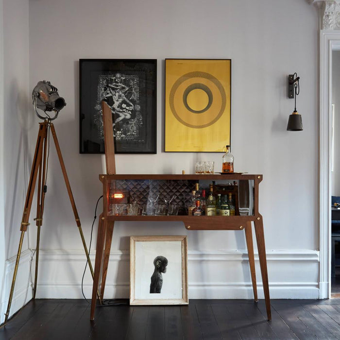 Rockstar Bar styled against a wall lined with spirits and glassware, with two paintings above, a wall sconce to the right, and an industrial floor lamp to the left.