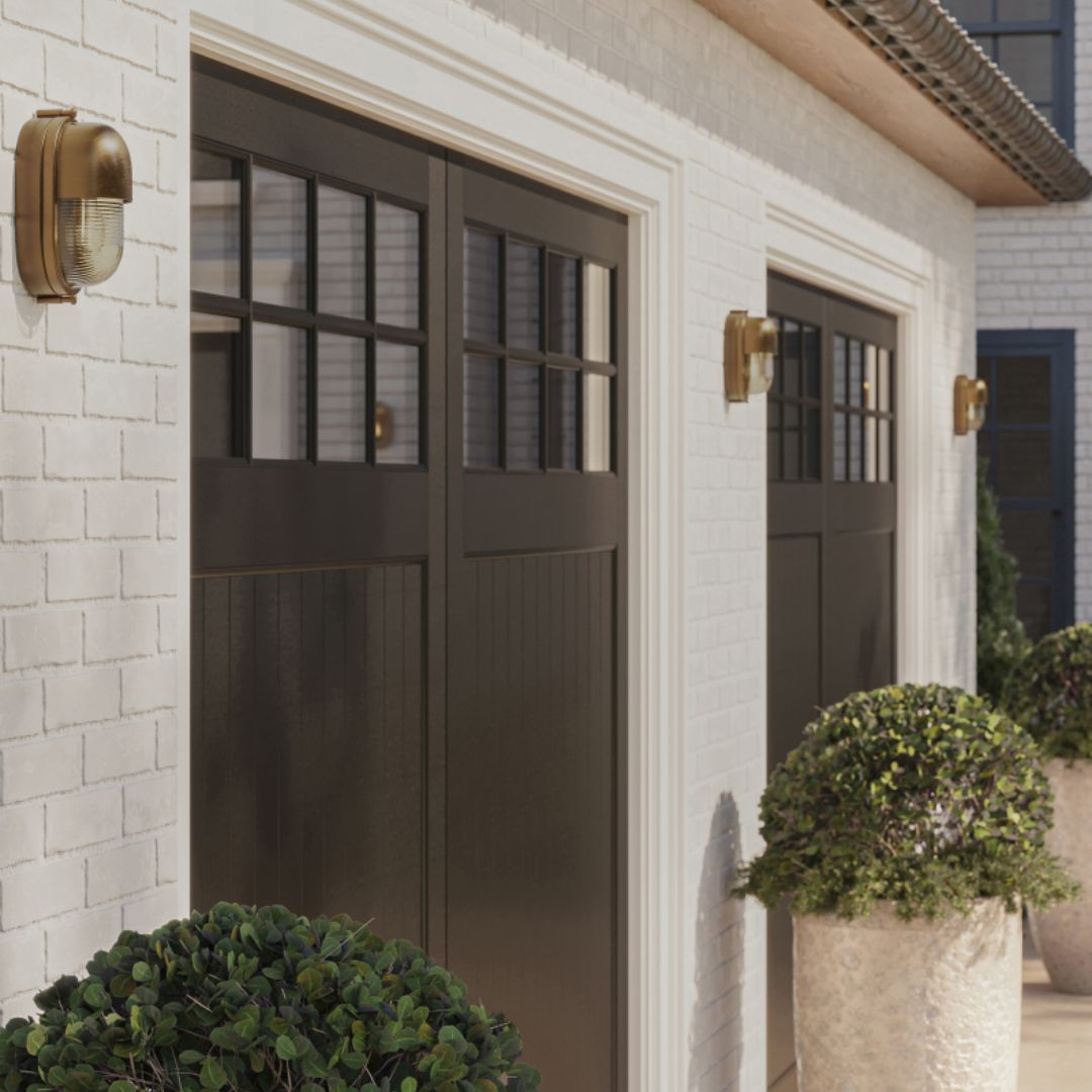 Three brass Maritime outdoor wall sconces mounted between two garage doors, set against white brick with black doors and greenery placed between them.