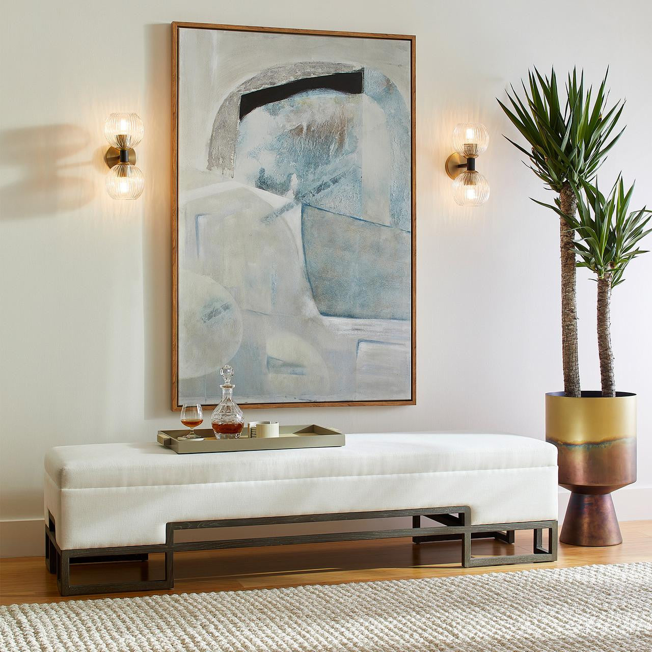 Vanora Bench styled in an entryway, accented by a potted plant on the left and a tray with a decanter on top, with two lit wall sconces framing a contemporary blue painting centered between them.