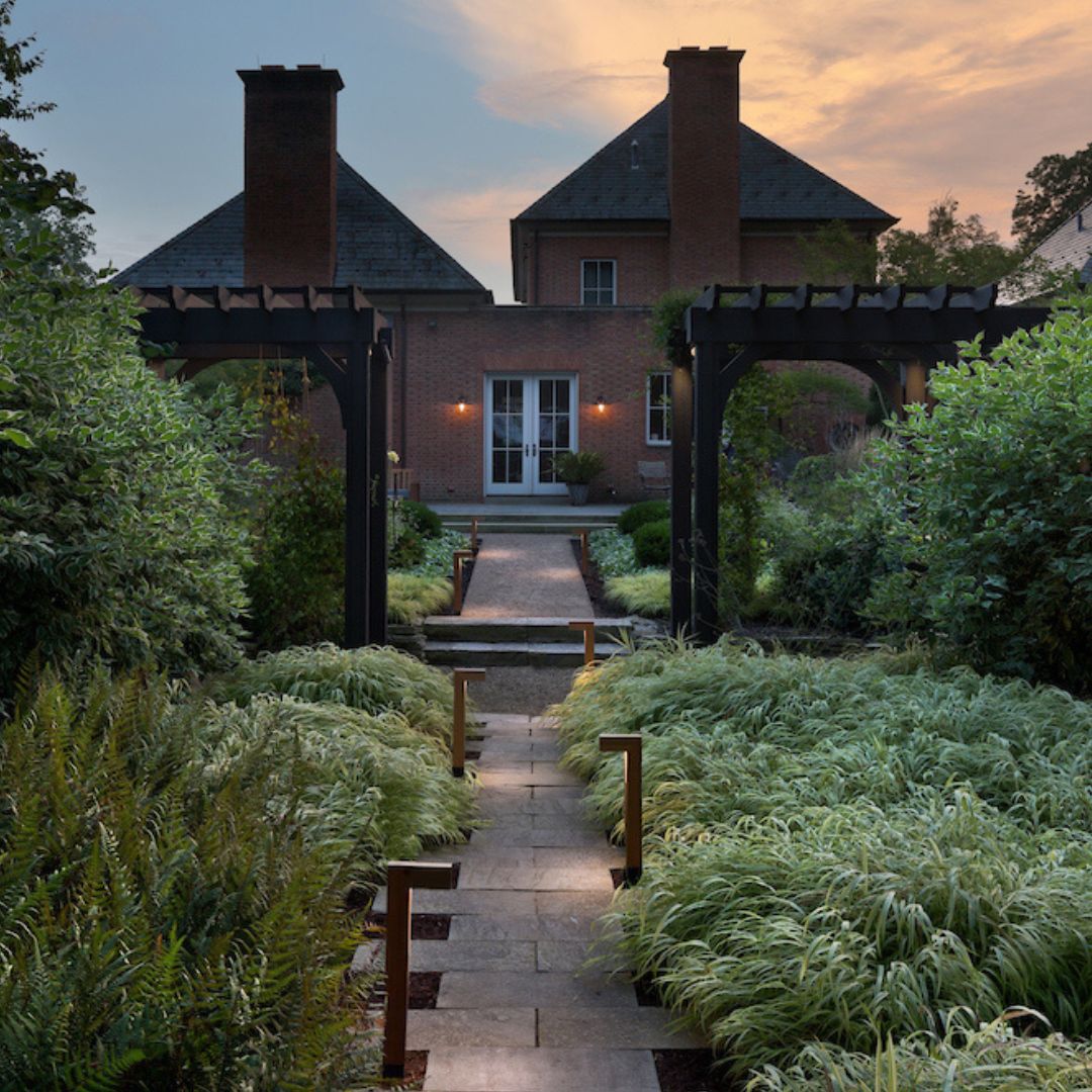 A grand backyard pathway illuminated with Atlantis path lighting and framed by lush greenery, leading toward a home with pergolas.