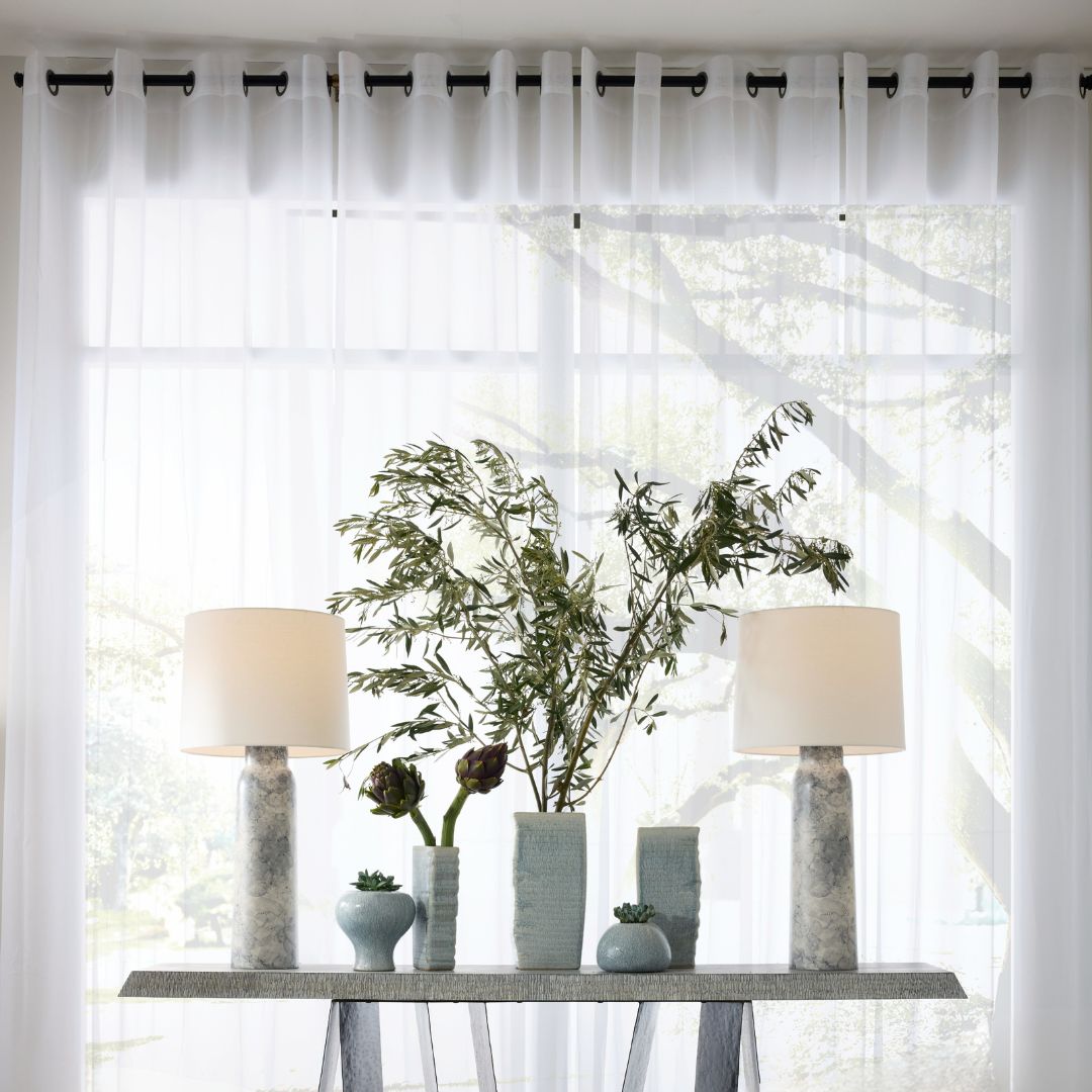 A pair of Heather Table Lamps arranged on a console table, accented with plants and vases, set in front of floor-to-ceiling windows with sheer curtains.