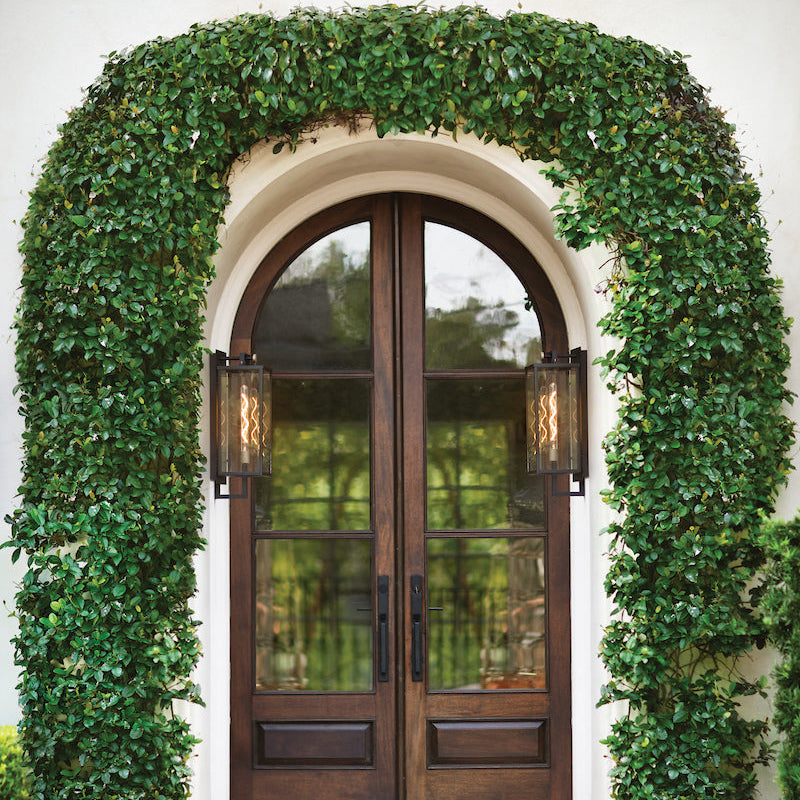 Wooden French door with glass panels framed by ivy on a white building with a Catalina Lantern flanking each side of the door.
