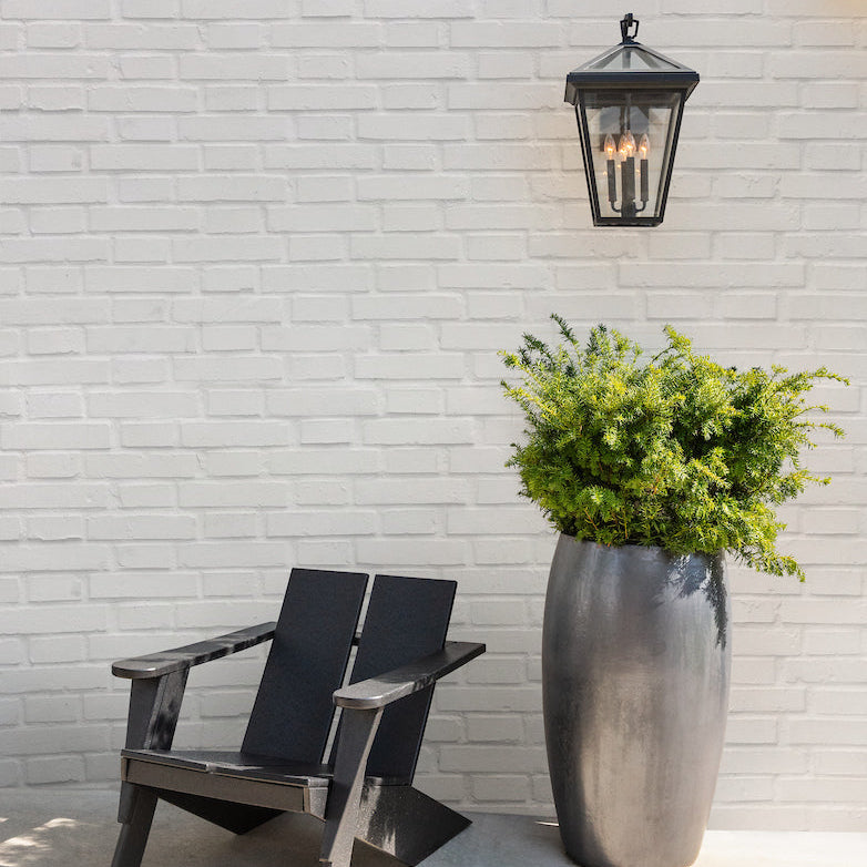 Outdoor setting with a wooden chair and potted plant against a white brick wall with the Alford Place Lantern installed.