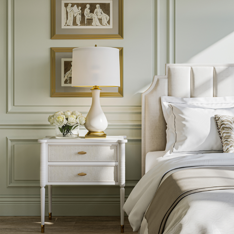 Neatly made bed with crisp white bedding, an ivory nightstand, a matching table lamp with antique gold accents, and coordinated decorative pillows in a serene bedroom setting.