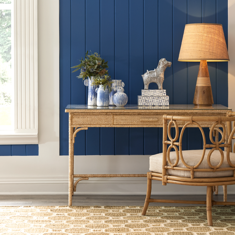 A coastal styled desk with a lamp and decorative items against a blue paneled wall.