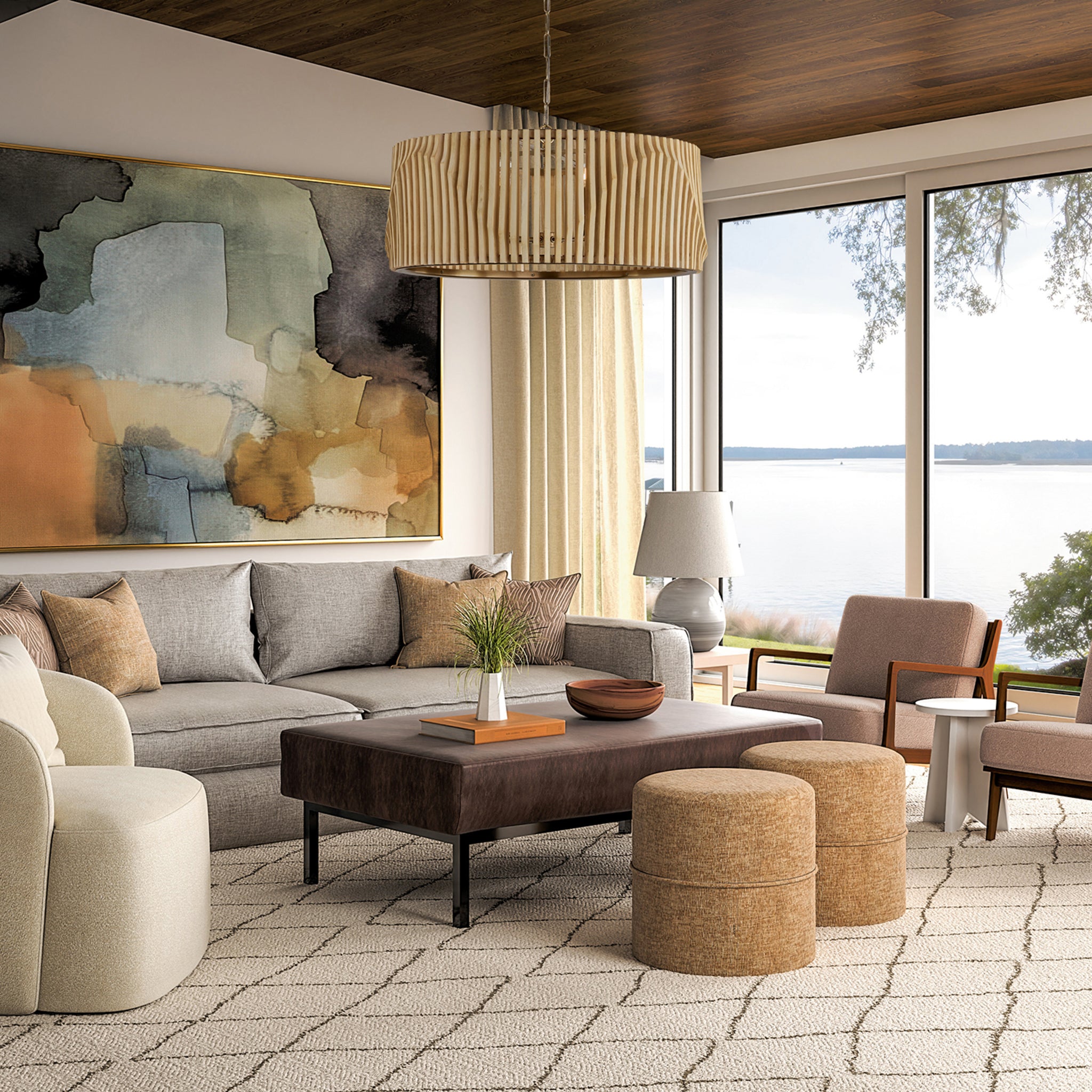 Open living room with gray sectional, leather ottoman, and tan poufs on a trellis rug, facing floor-to-ceiling lake views with the Capital Archer 6-Light Pendant in Light Wood and Matte Black styled in the center.