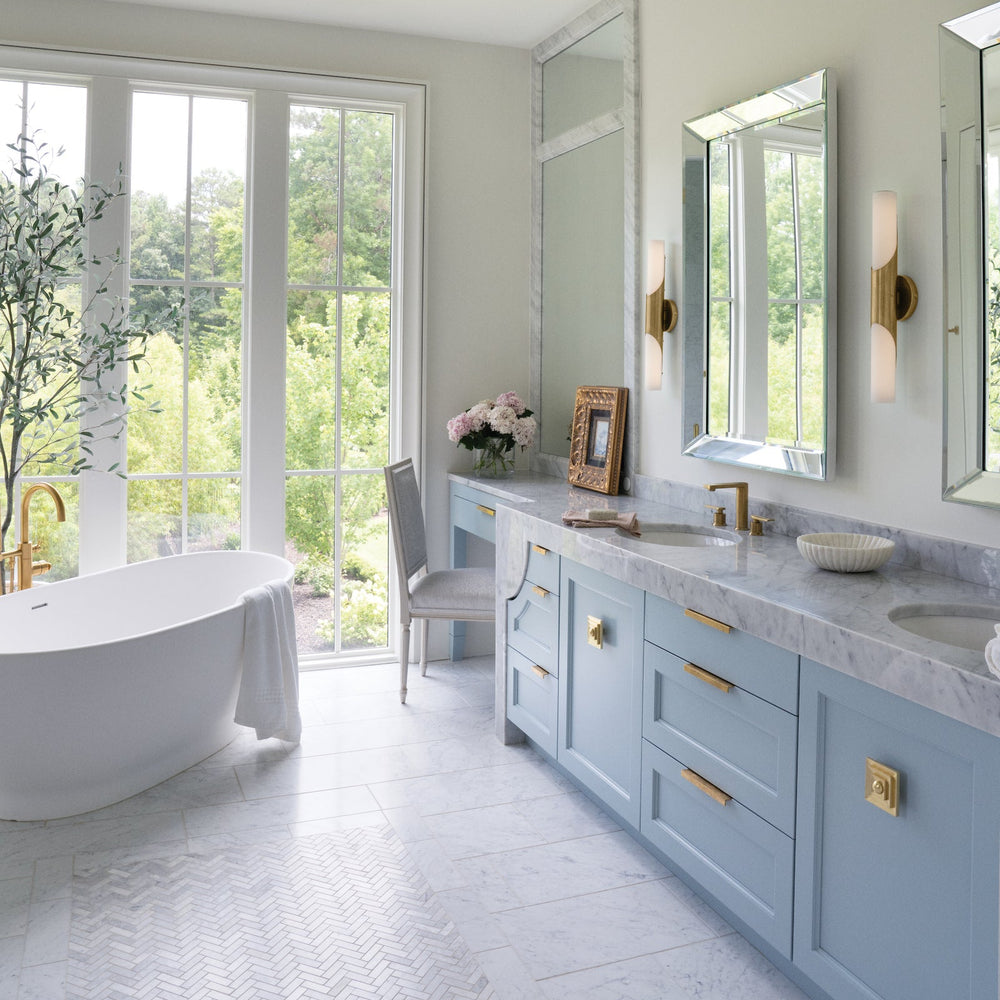 Modern bathroom with light blue vanity, white freestanding bathtub, and large windows.