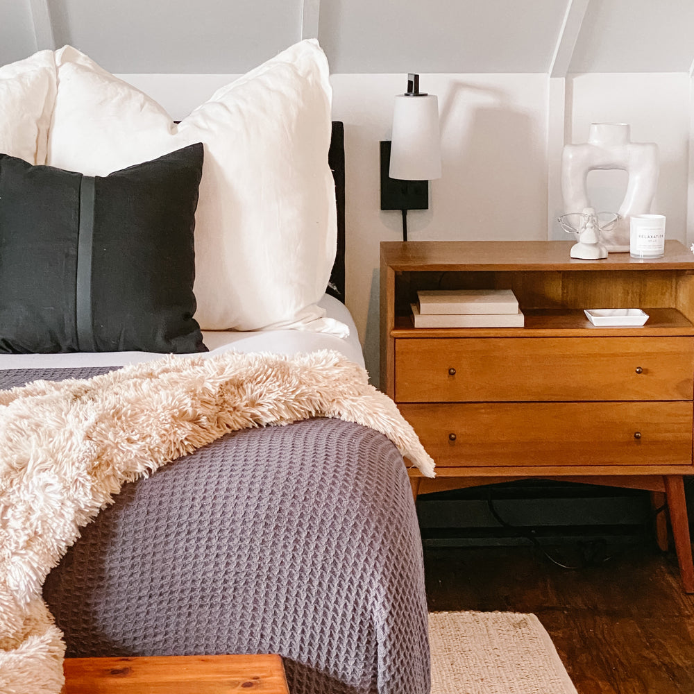Bedroom with a bed featuring pillows and a blanket, next to a wooden nightstand with books and wall sconce.