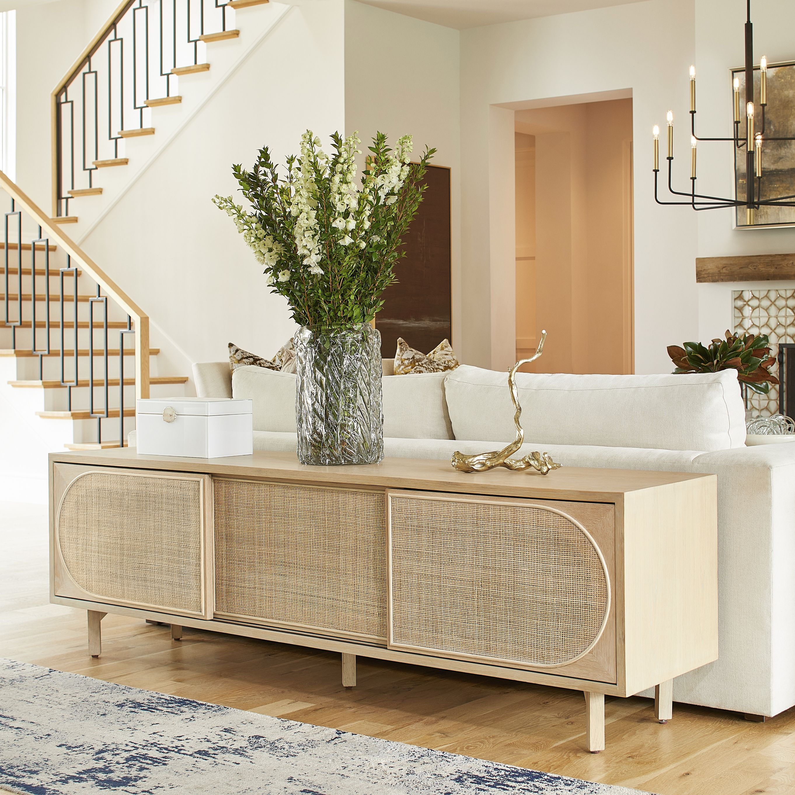 Styled behind an ivory sofa in a living room, the Lamu Media Cabinet features a contemporary sculpture on the right, a clear vase filled with lush green and ivory blooms at the center, and a refined ivory box positioned to the left.