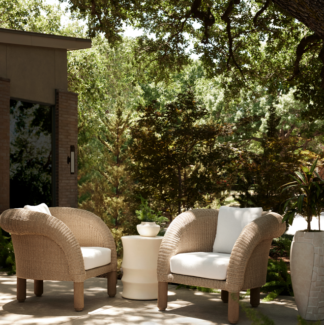 Two wicker chairs with white cushions on a patio under a tree