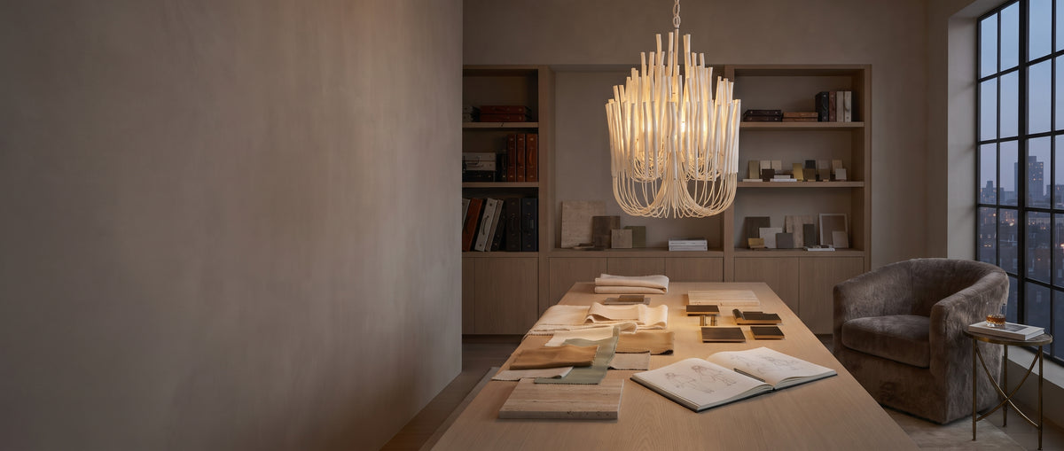 Tilda chandelier in white hanging over an oak worktable styled with fabric swatches, finish samples and an open sketchbook in an elegant interior design studio at dusk.