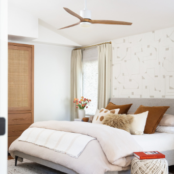 A warm transitional bedroom with a bed, ottoman, a wood and white ceiling fan styled above the bed.