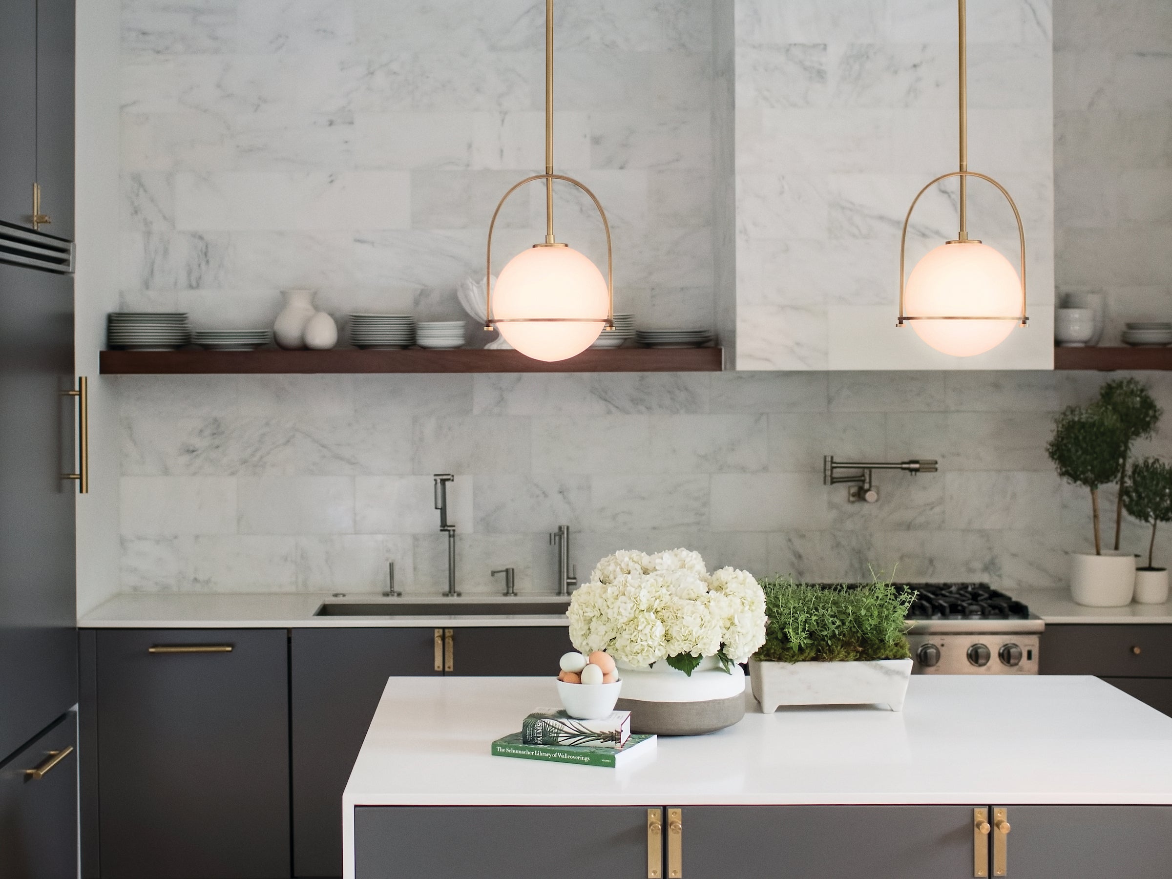 Modern kitchen with gray marble backsplash, gray cabinetry with brass hardware, two brass pendant lights with milk glass, and white countertops.