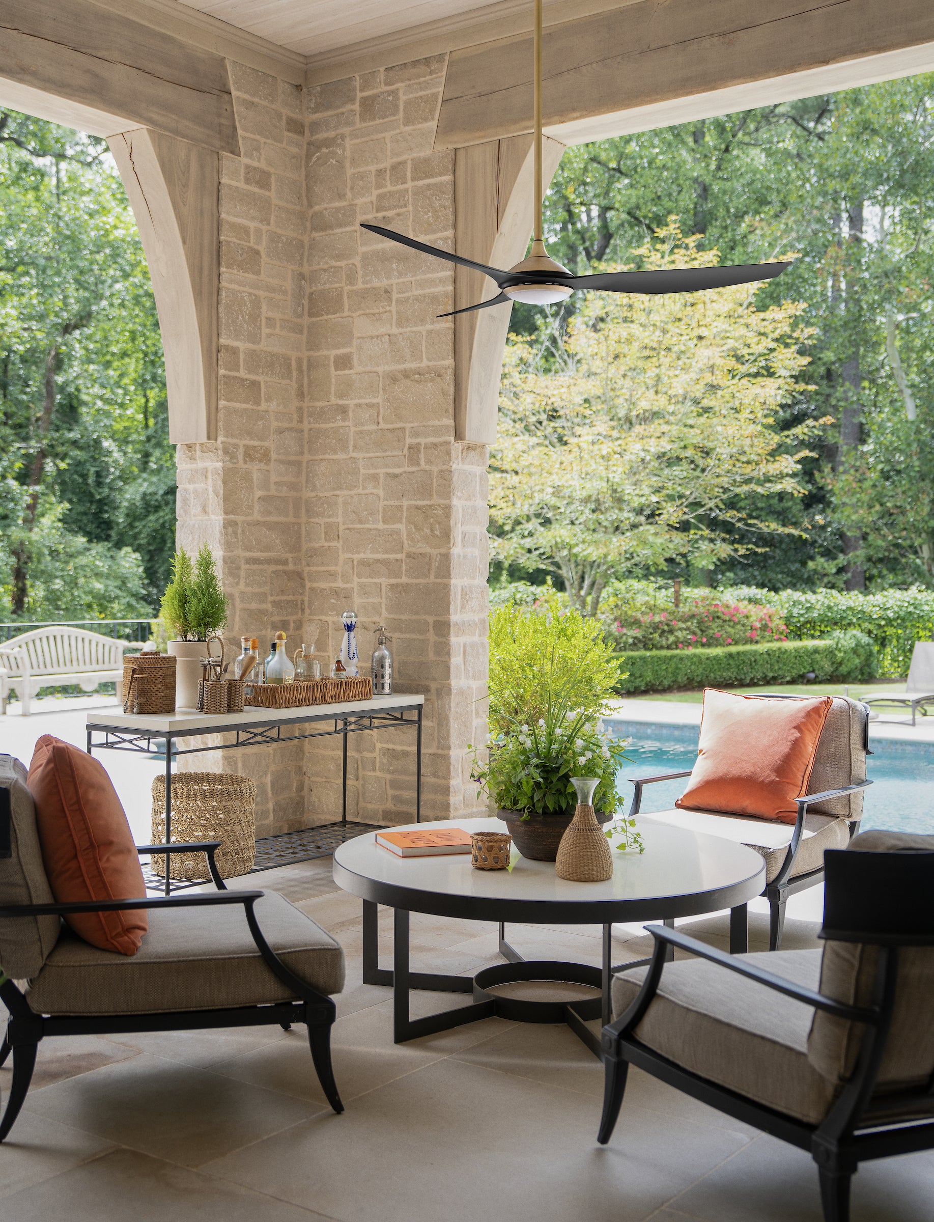 Outdoor patio furniture set featuring a round white coffee table with a black metal base, surrounded by three gray upholstered armchairs with black frames and rust-orange pillows, under a ceiling fan on a stone-pillared porch overlooking a pool and green garden.