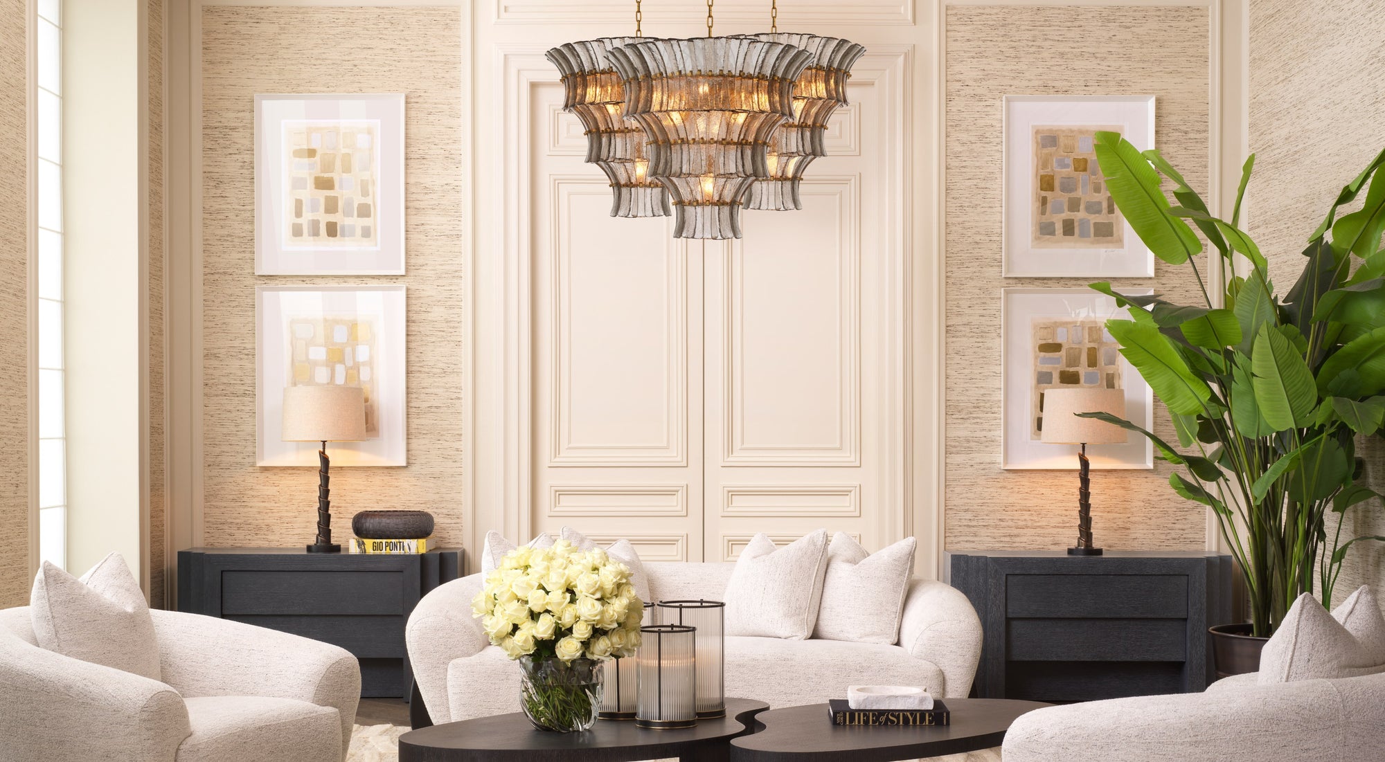 A luxury modern chandelier with a woven metal cage and warm glowing bulbs, hanging in a beautifully styled living room with beige walls, a white sofa, and a large green plant.