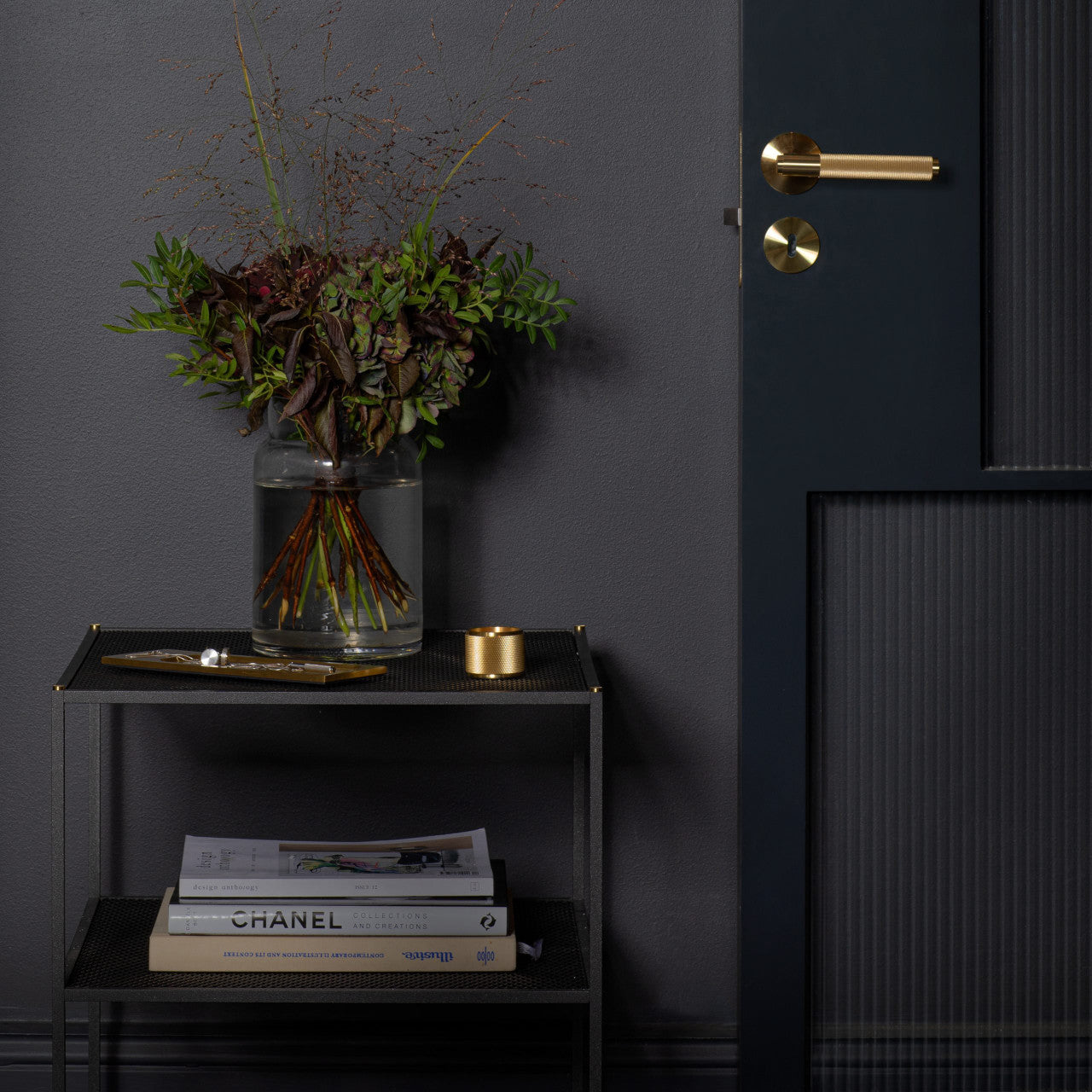 Dark, moody entryway with a brass door lever, foliage arrangement in a glass jar, and stacked design books on the Buster + Punch Meshed Side Table.